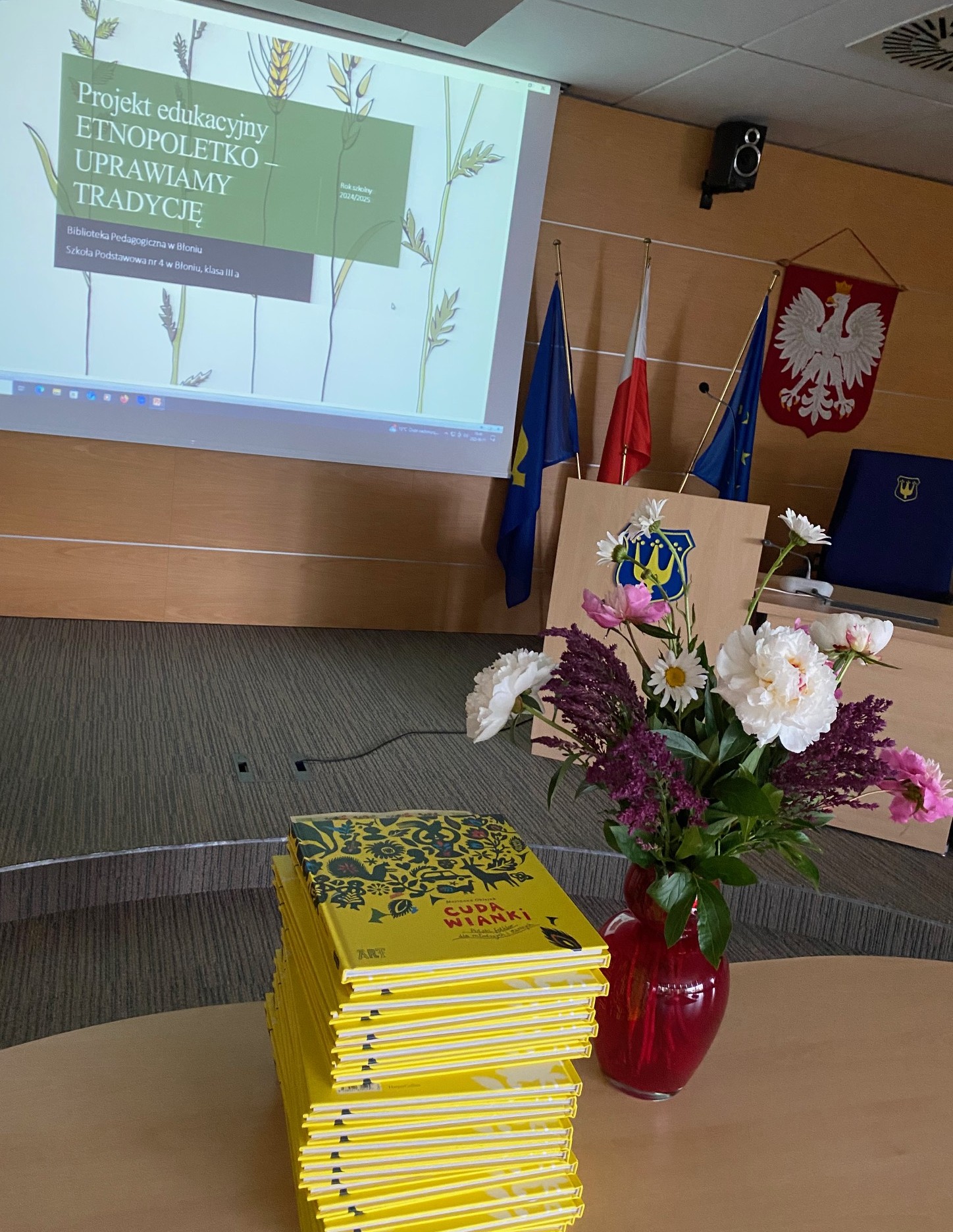Na pierwszym planie na stole stoi Iwazon z kwiatami oraz stos książek, w tle ekran z wyświetlonym napisem ,,Projekt edukacyjny ETNOPOLETKO - UPRAWIAMY TRADYCJĘ"
