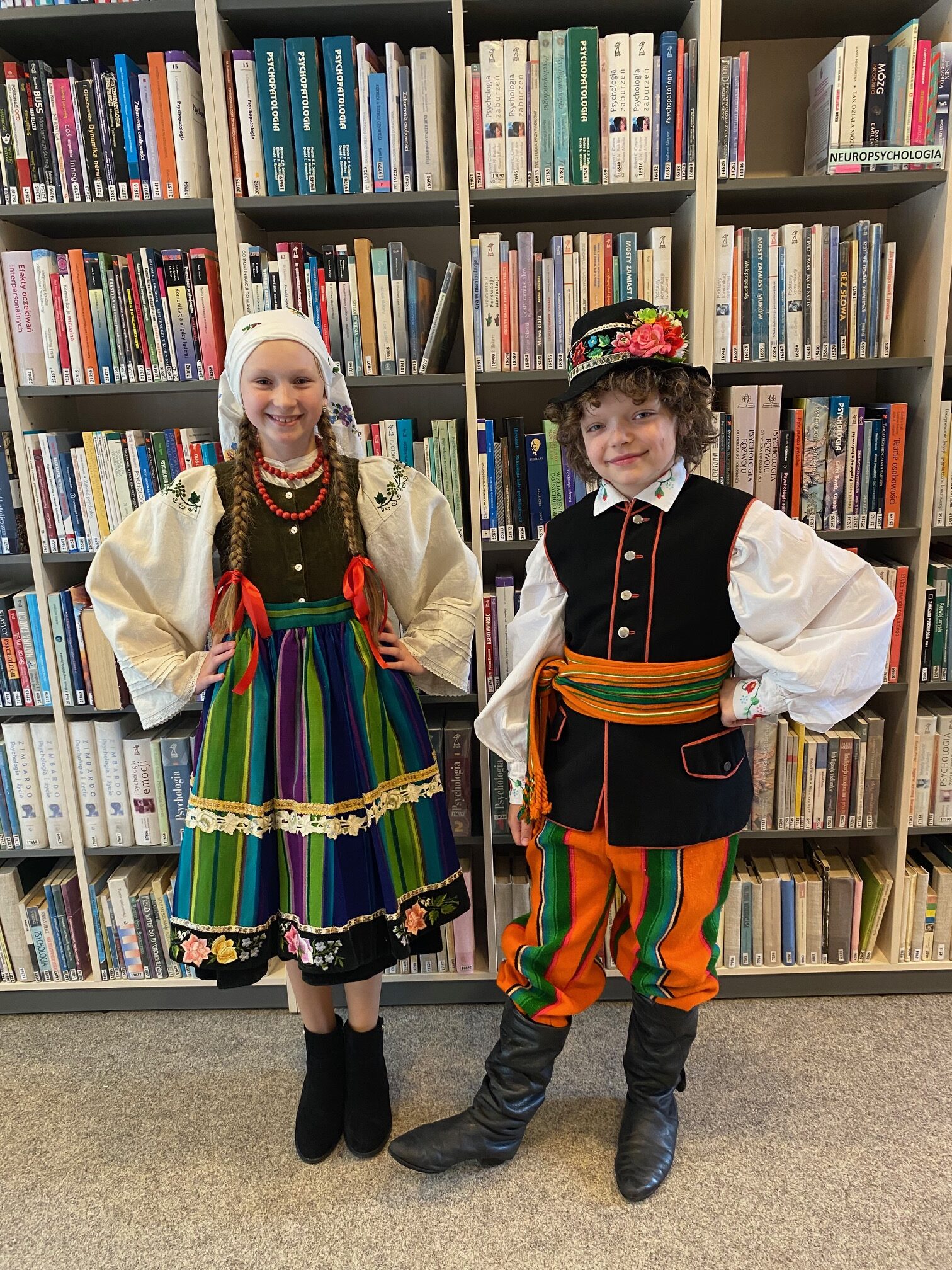 Dwoje dzieci w łowickich strojach ludowych na tle regału z książkami