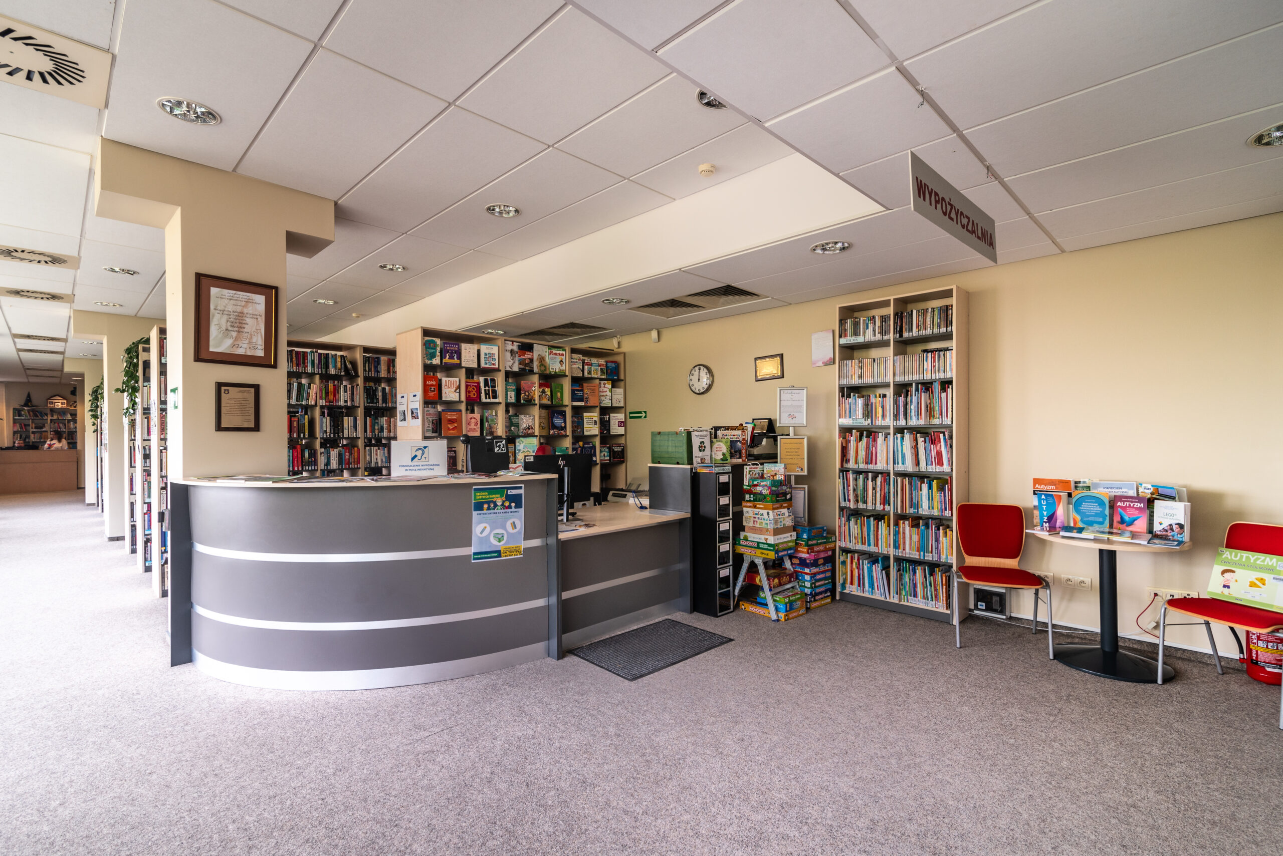 Jasne i przestronne wnętrze biblioteki w Błoniu. Na pierwszym planie znajduje się nowoczesna, łukowata lada biblioteczna w kolorze szarym z jasnym blatem. Za ladą widoczne są regały wypełnione kolorowymi książkami dla dzieci i dorosłych. Po prawej stronie, przy ścianie, stoi stolik z ekspozycją książek oraz dwa czerwone krzesła. Na suficie zamontowane są estetyczne kasetony i punktowe oświetlenie